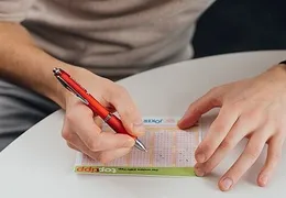 Person filling out a lottery ticket with a red pen at a round white table, wearing a smartwatch on their left wrist.