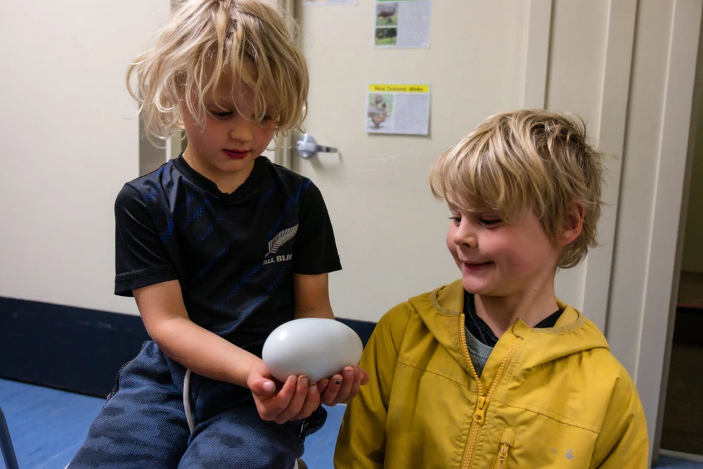 Two young children indoors, one holding a large, oval-shaped kiwi egg