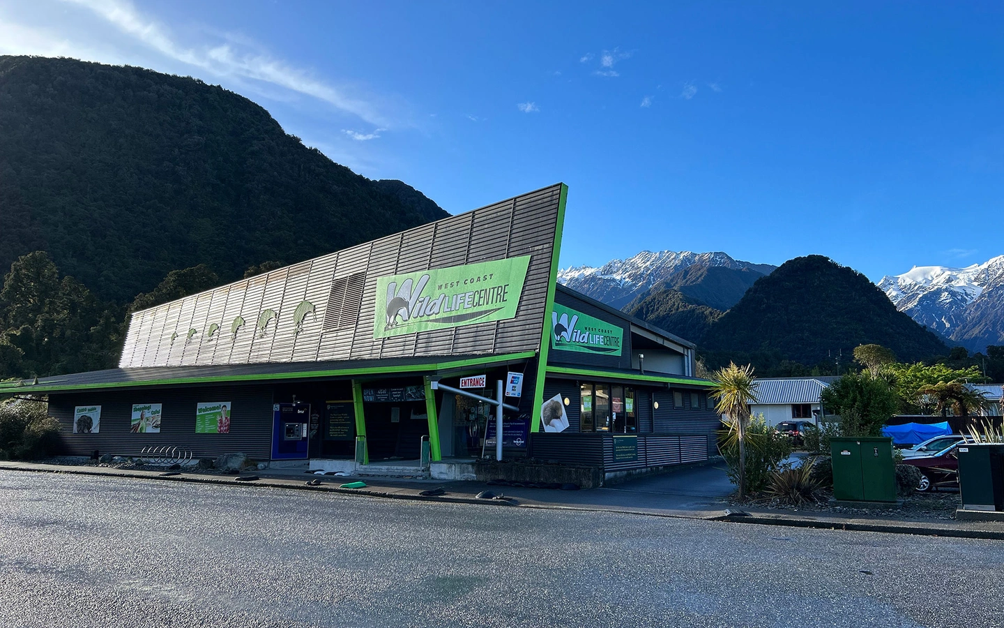 A modern wildlife centre building, set against a backdrop of mountains and a clear blue sky.