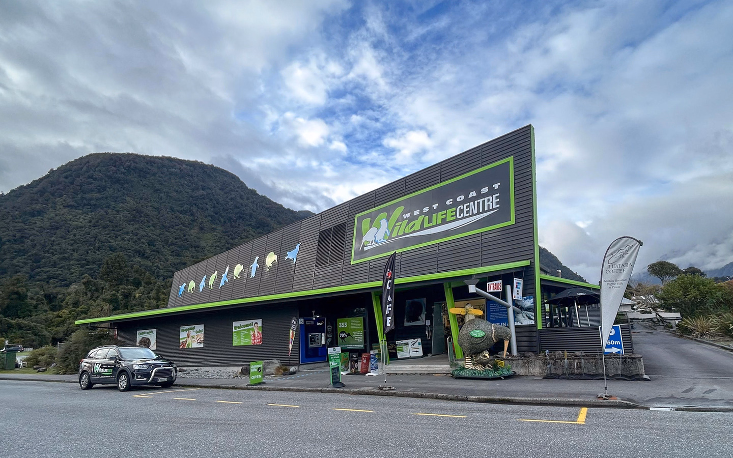 the West Coast Wildlife Centre front building with a parked car, surrounded by mountains and a cloudy sky.