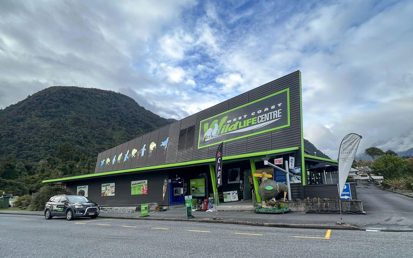 the West Coast Wildlife Centre front building with a parked car, surrounded by mountains and a cloudy sky.