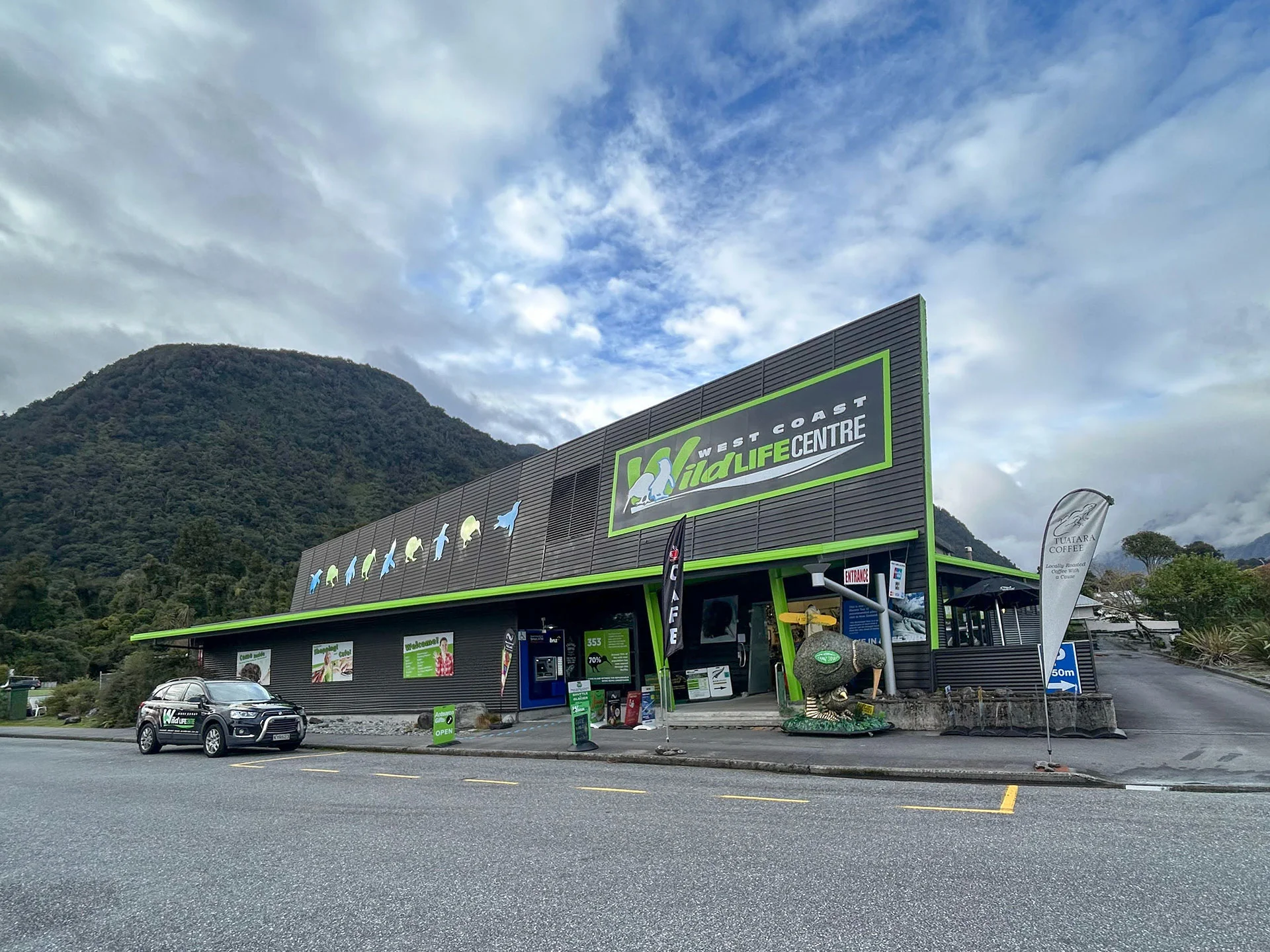 the West Coast Wildlife Centre front building with a parked car, surrounded by mountains and a cloudy sky.