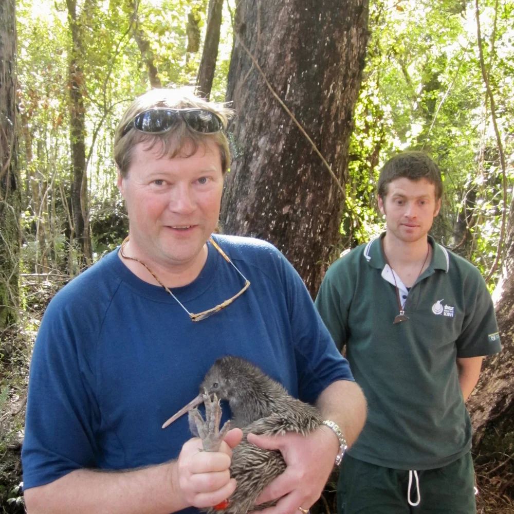Two people in a forest, one holding a kiwi bird. Sunlight filters through the trees, illuminating the scene.