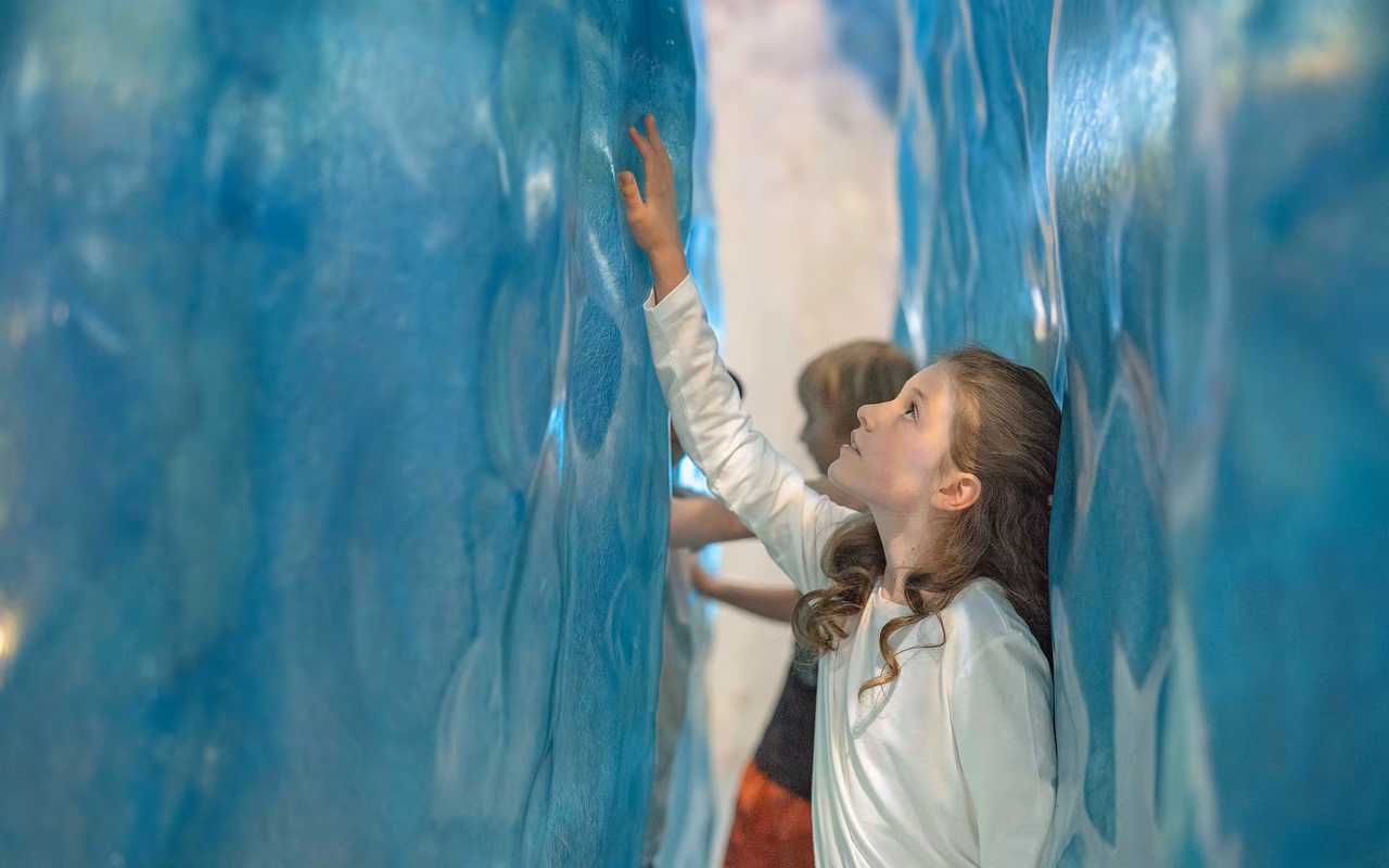 A girl in a white shirt touches a blue, textured blue wall, looking up thoughtfully
