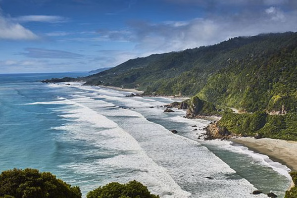 Scenic view of a coastal landscape with waves crashing on the shore, lush green hills, and a partly cloudy sky.