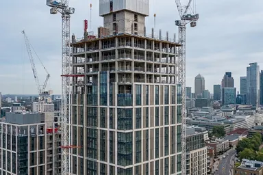Skyscraper under construction with cranes, surrounded by cityscape featuring modern high-rise buildings and a cloudy sky.