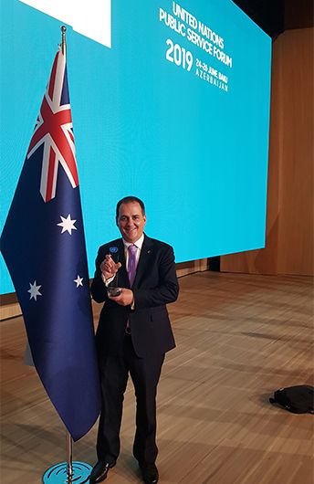 Andrew Woolnough holding his United Nations Public Service Award beside an Australian flag at the 2019 UN Public Service Forum in Baku