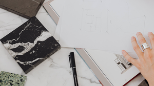 A hand with a silver ring holds a sketch on paper, surrounded by marble samples and a pen on a marble surface.