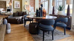 Modern living room with a brown sofa, round wooden table, dark chairs, and decorative vases. Large windows provide natural light.