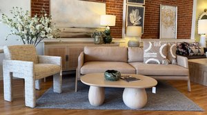 Modern living room with a tan sofa, patterned cushions, textured chair, round wooden coffee table, art on brick wall, and a floral arrangement.