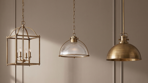Three pendant lights in brass finish hang against a beige wall; one with a lantern shape, one with a glass dome, and another with a solid dome.