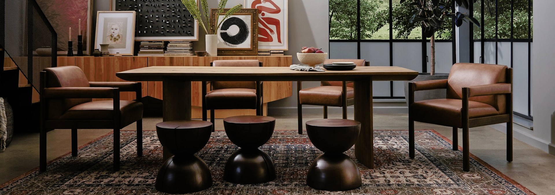 Modern dining room with a wooden table, leather chairs, round stools, art pieces, and large windows with views of greenery.