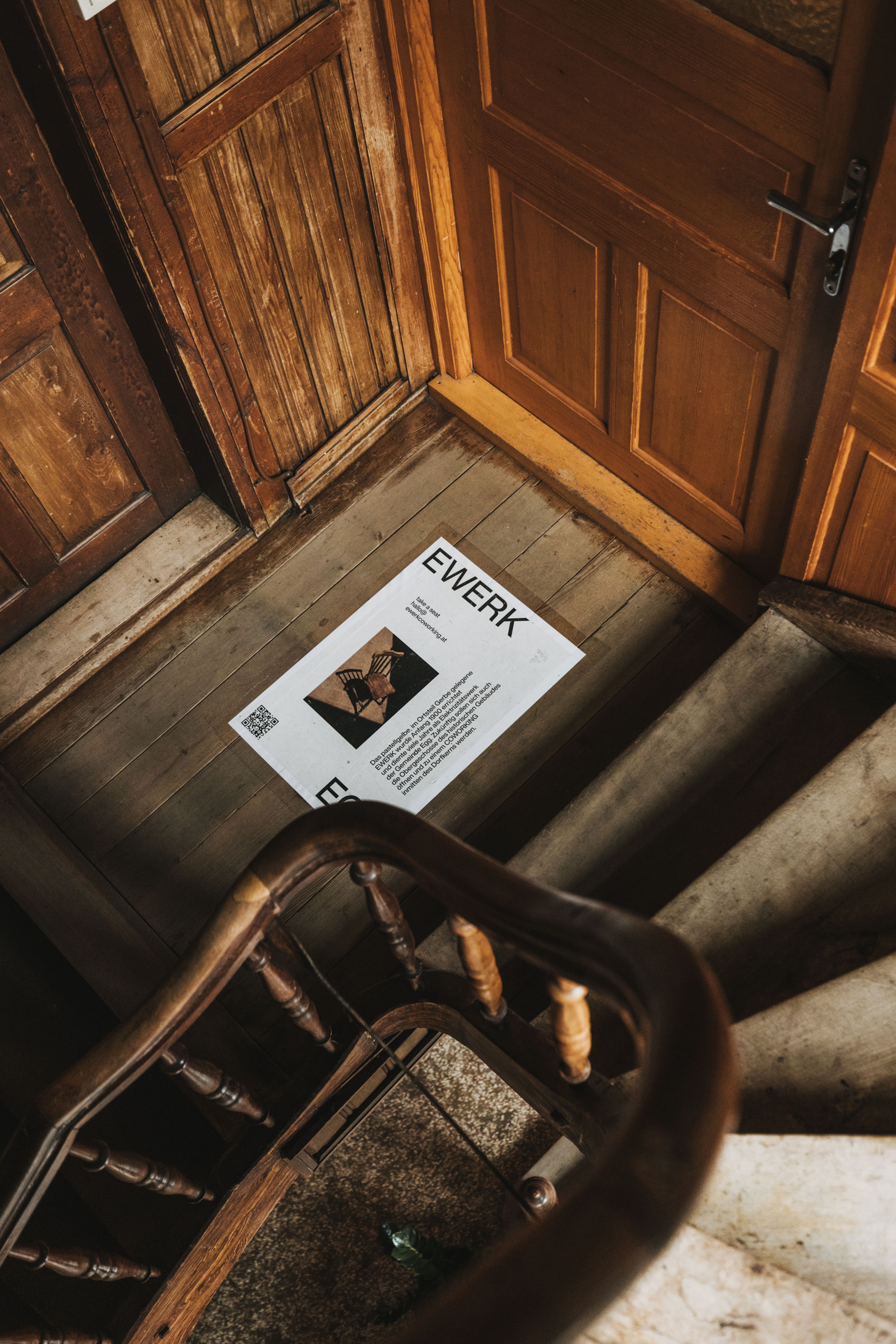 Poster mit dem Titel „EWERK“ auf dem Boden eines Treppenabsatzes aus Holz, umgeben von Holztüren und Geländern.