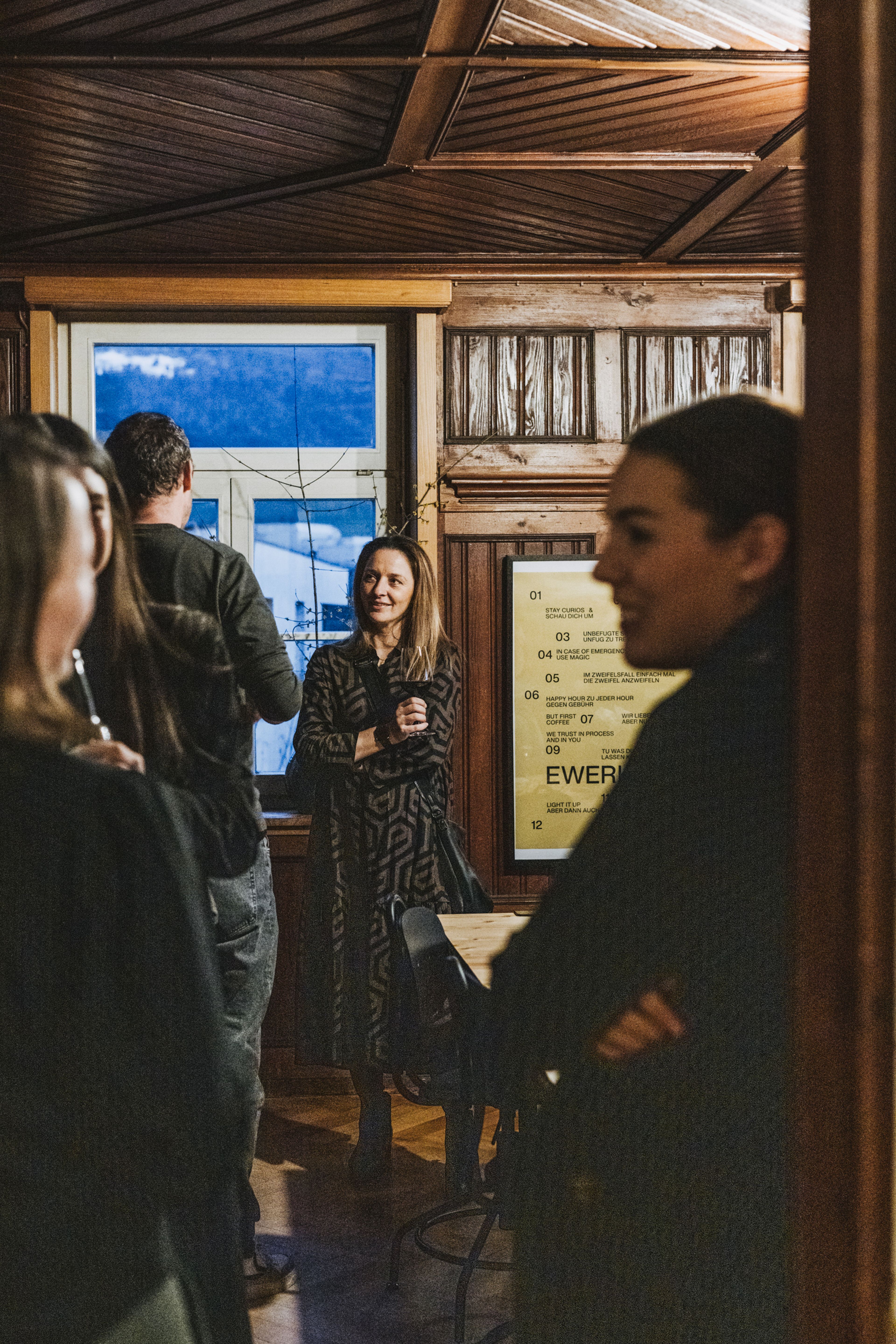 Eine Gruppe von Menschen unterhält sich in einem warm beleuchteten Raum mit Holzwänden und einem Fenster, durch das man eine verschneite Landschaft sehen kann.