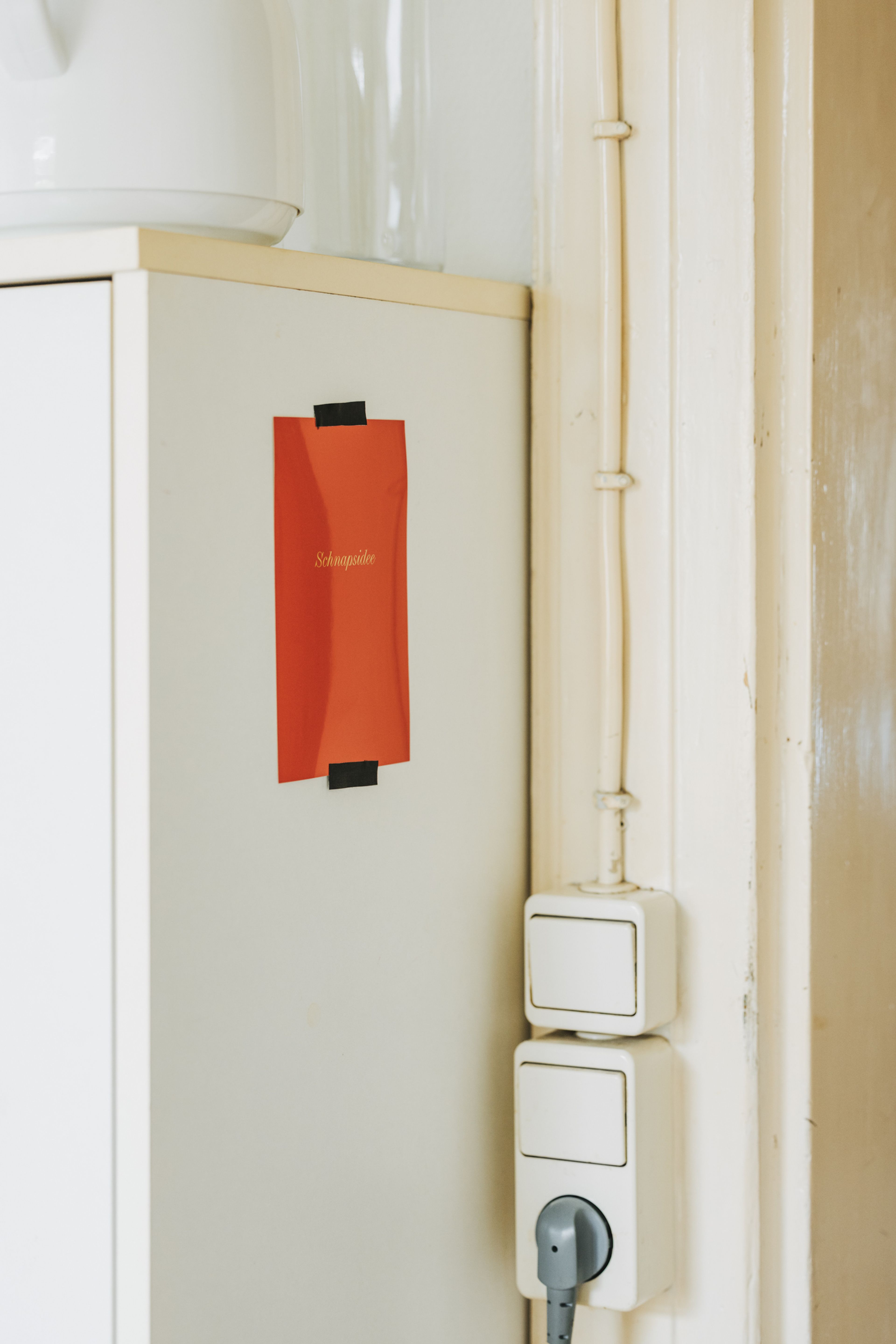 Rotes Papier mit der Aufschrift „Schokolade“ auf einem Schrank in der Nähe eines Lichtschalters und eines eingesteckten Kabels, in einem Raum mit beigen Wänden.