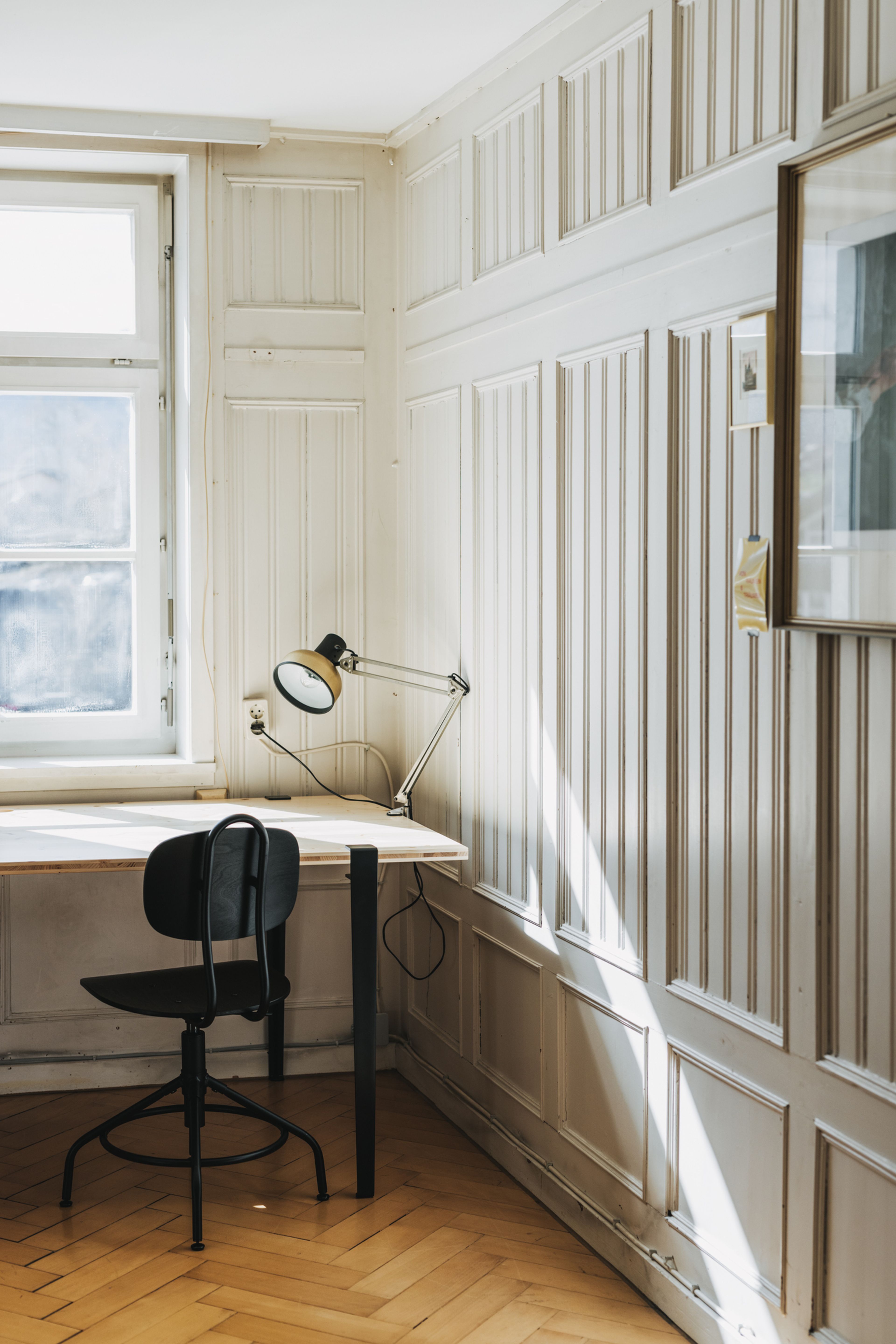 Minimalistischer Arbeitsbereich mit einem Schreibtisch aus Holz, einem schwarzen Stuhl und einer verstellbaren Lampe am Fenster. Sonnenlicht fällt herein und hebt den Fischgrätenboden hervor.