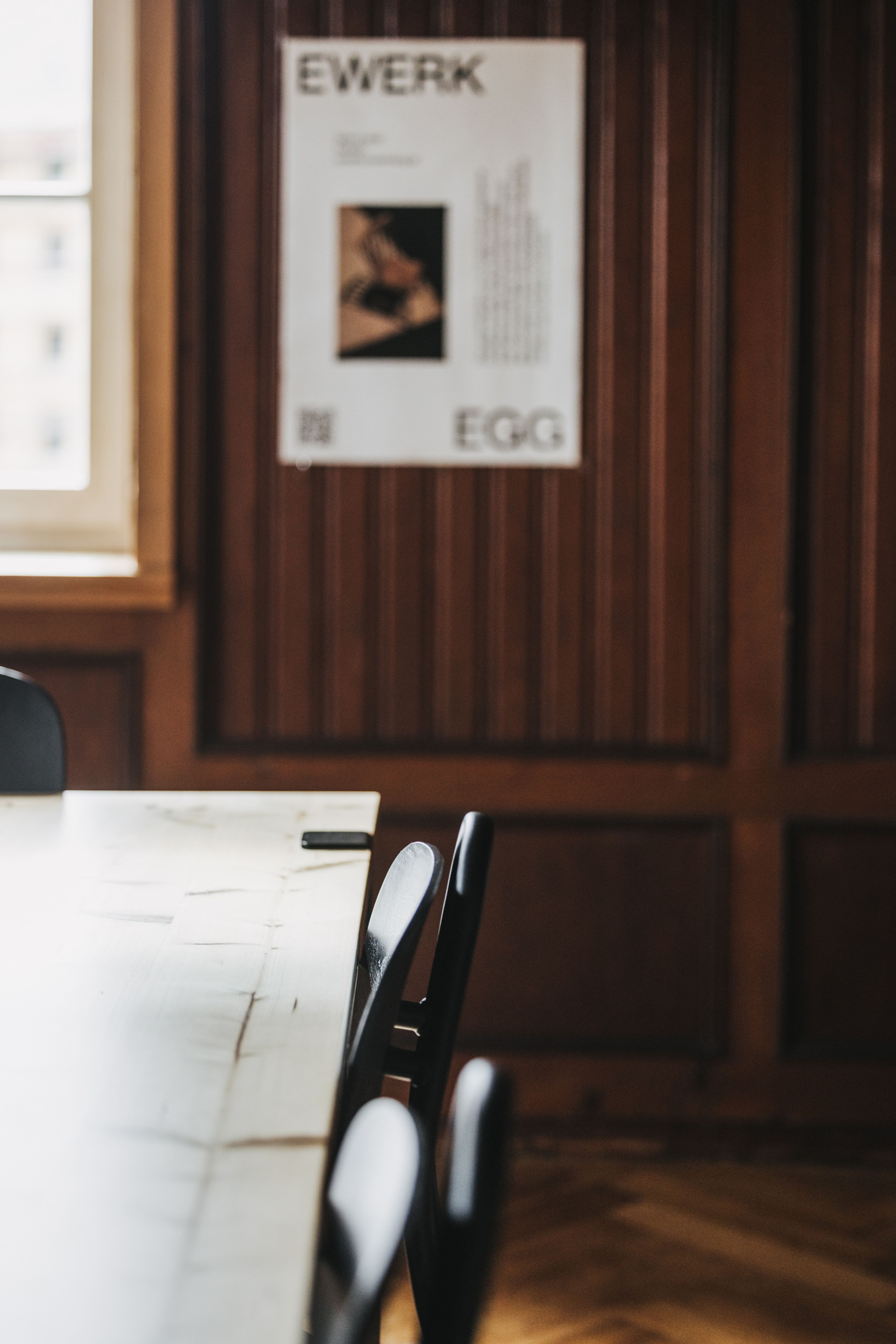Holzgetäfelter Raum mit einem Tisch und Stühlen. An der Wand hängt ein Poster, das teilweise sichtbar ist und Text sowie ein Bild enthält.