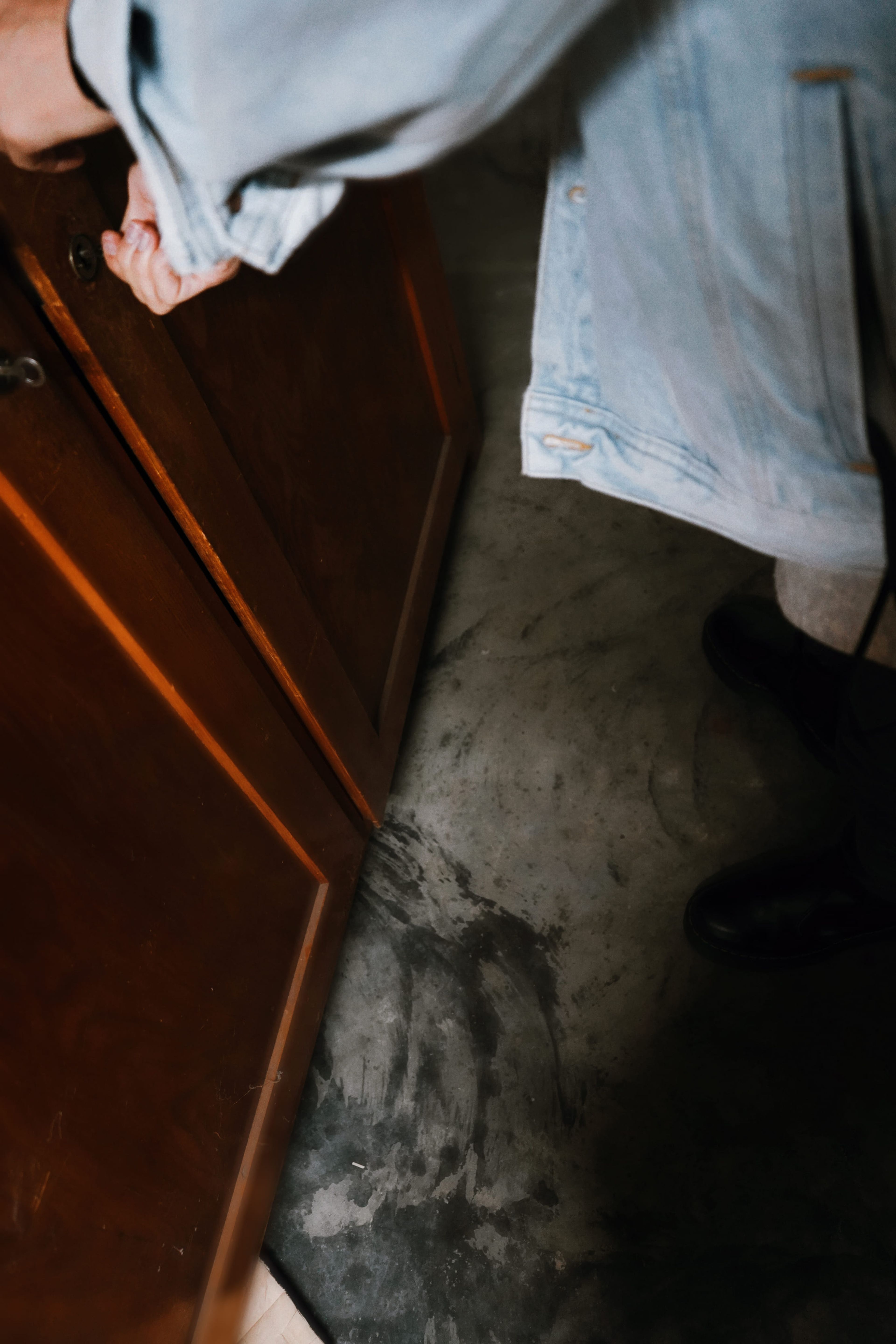 Person in einer leichten Jacke öffnet eine Holzschranktür und steht auf einem Betonboden mit sichtbaren Schuhabdrücken.
