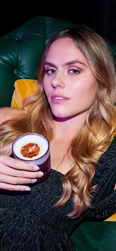 Junge Frau, die auf einem grünen Ledersofa liegt, einen schaumigen Violet Sour mit einer getrockneten Zitrusscheibe in der Hand hält und in die Kamera blickt.