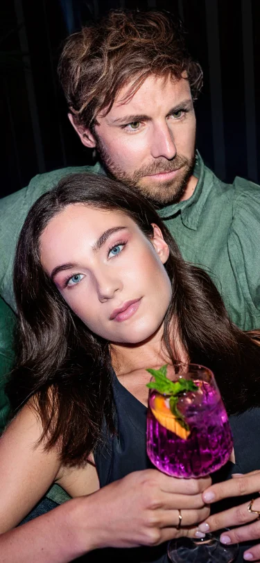 Ein Paar sitzt entspannt; die Frau lehnt sich an den Mann, sie hält einen Violet Spritz mit Orangenscheiben und Minze in der Hand.