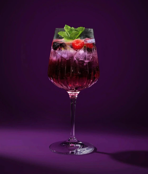 Weinglas mit einem Violet Wild Berry, Eiswürfeln, Himbeeren, Blaubeeren und Minzgarnitur vor einem tiefvioletten Hintergrund.
