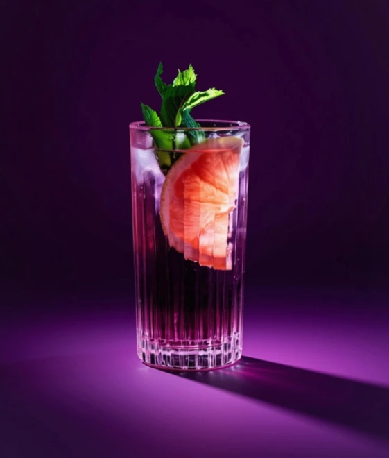 Highball-Glas mit Violet Tonic, Eis, Grapefruitscheibe und Minzezweig, violett beleuchtet vor passendem Hintergrund.