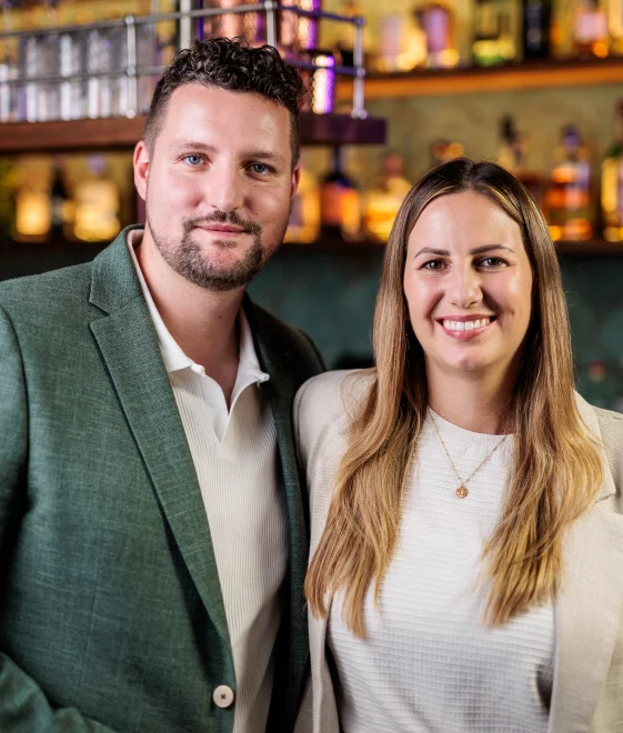 Lächelnder Mann und Frau in schicker Freizeitkleidung posieren zusammen in einer warm beleuchteten Bar mit Flaschen in Regalen hinter ihnen.