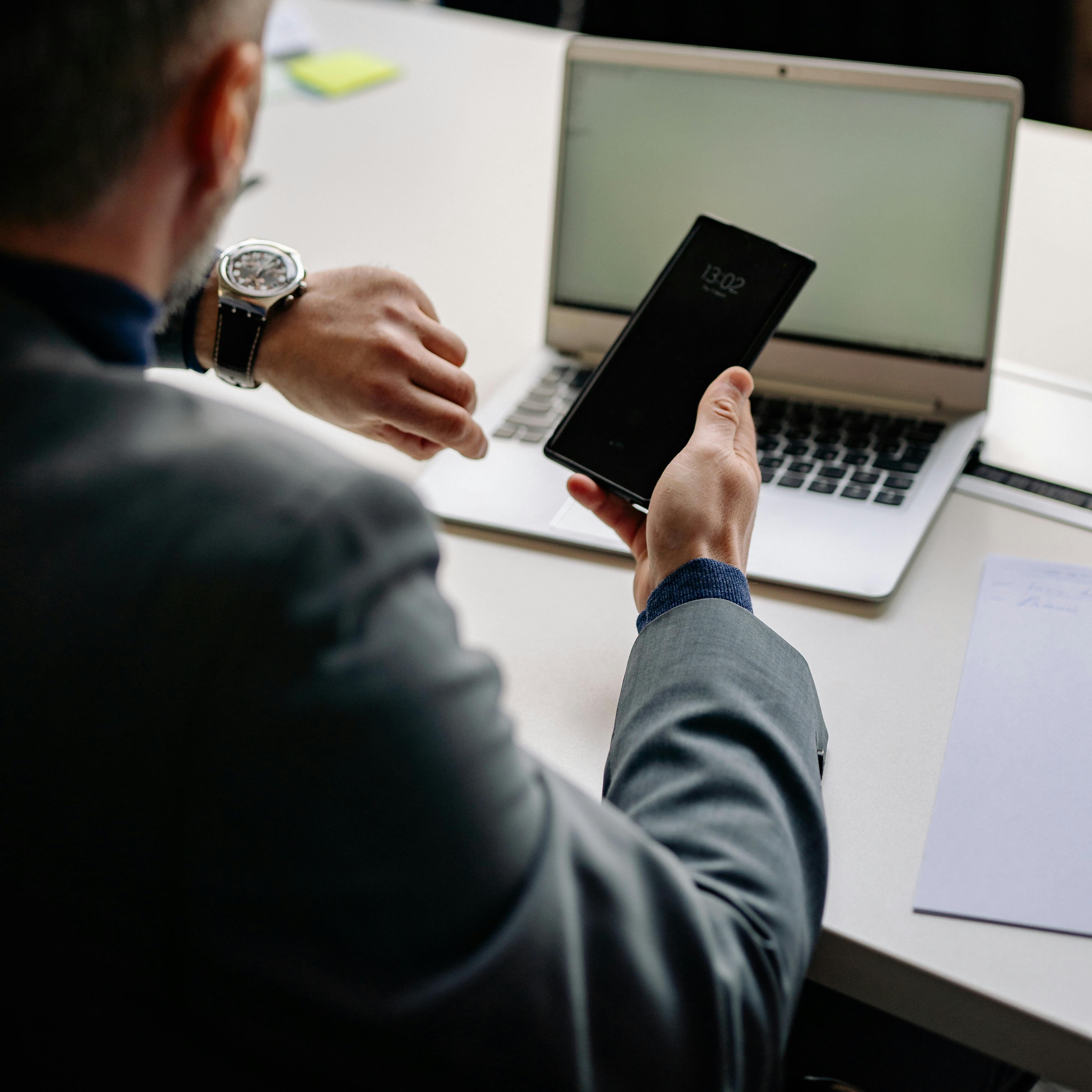 Person in a suit holding a smartphone, seated at a desk with an open laptop and a sheet of paper.
