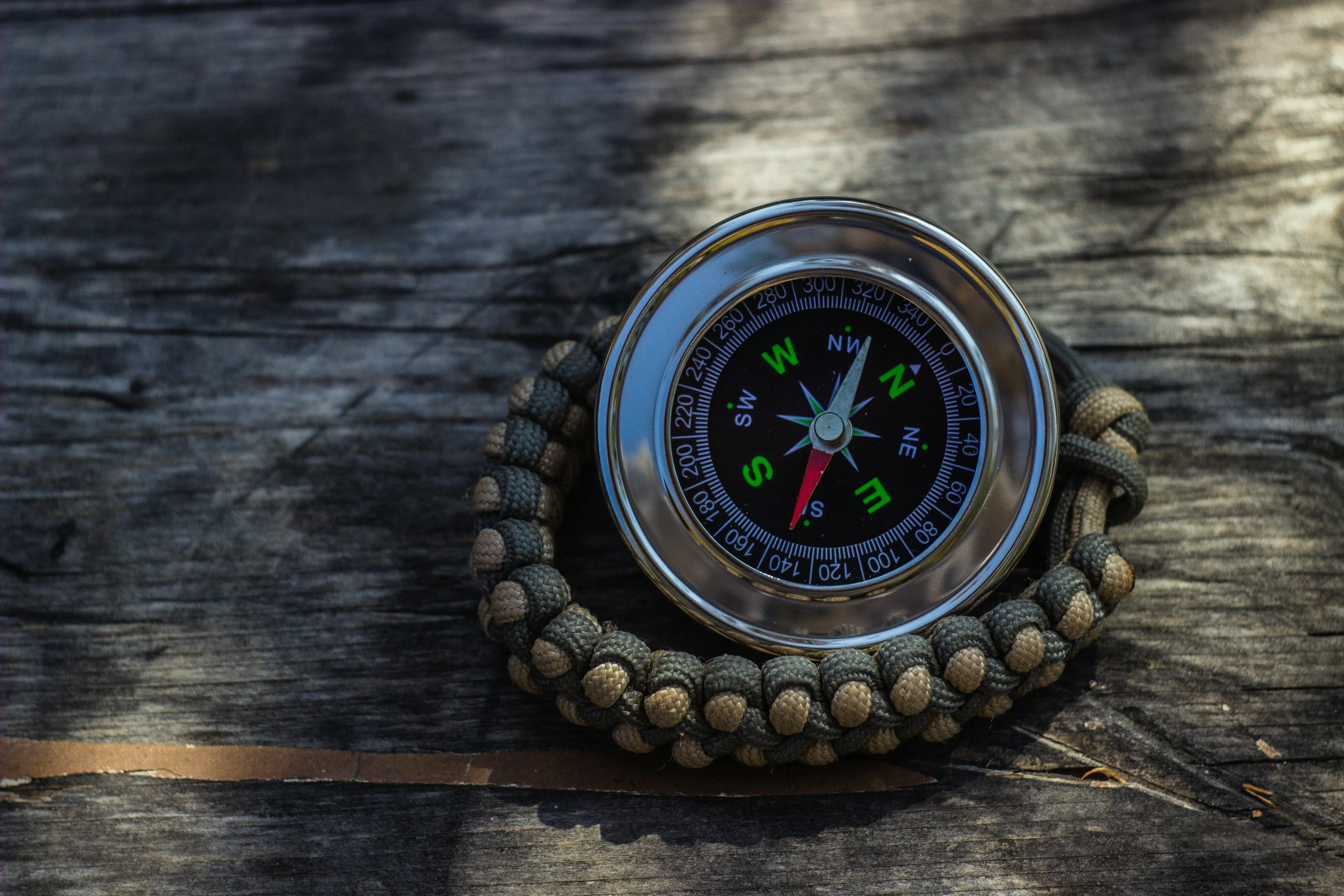 A compass with a black face and colorful directions rests on a braided rope bracelet on a weathered wooden surface.