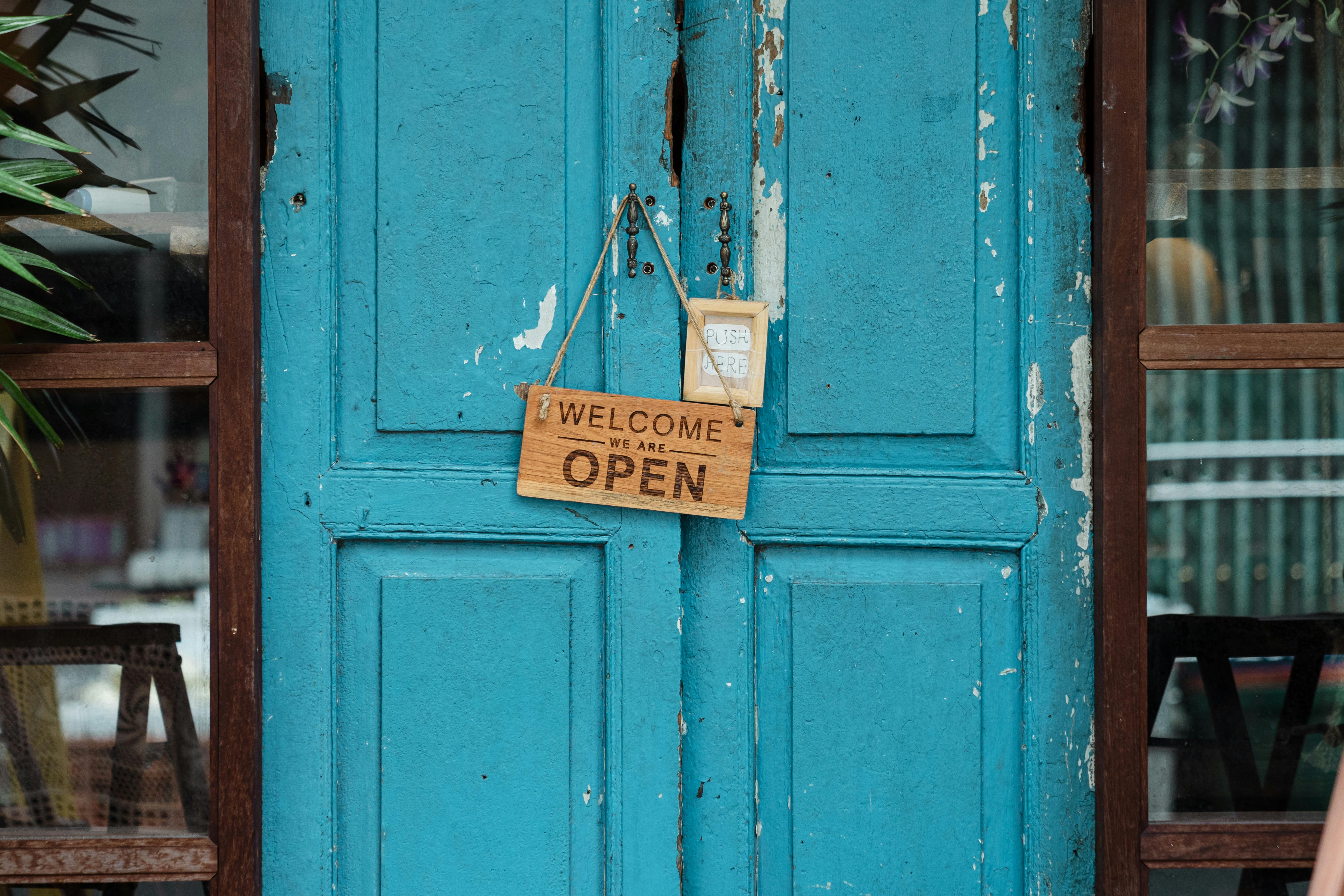 A rustic blue door with a wooden "Welcome, We Are Open" sign hanging on it. Flanked by glass panels and greenery.