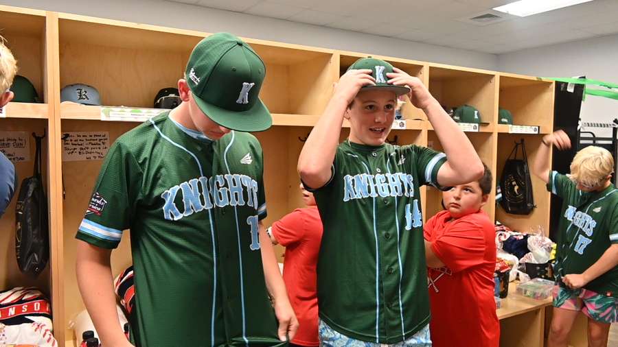 Young baseball players in green Knights uniforms trying on caps in a locker room.