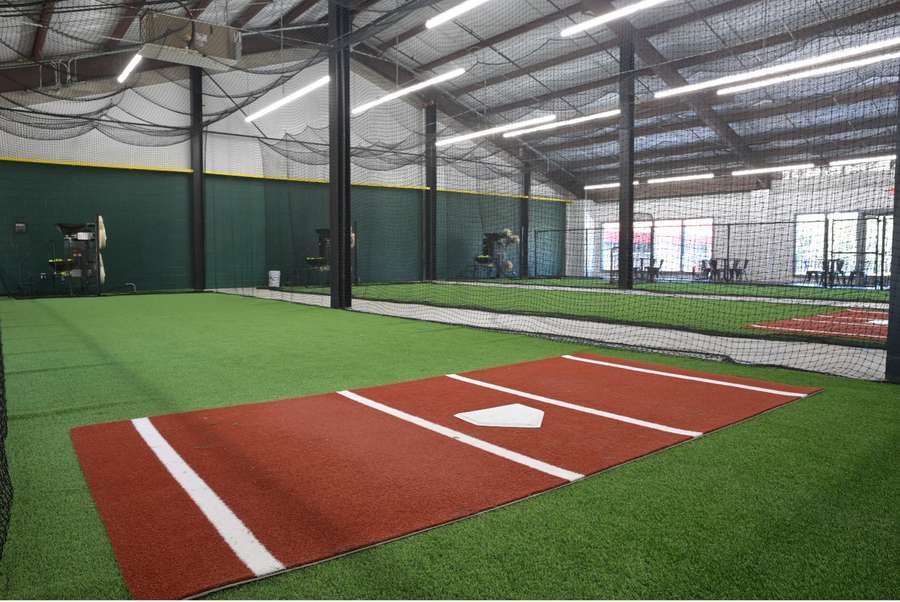Indoor baseball facility with multiple batting cages, green turf, and red clay diamonds under bright lighting.