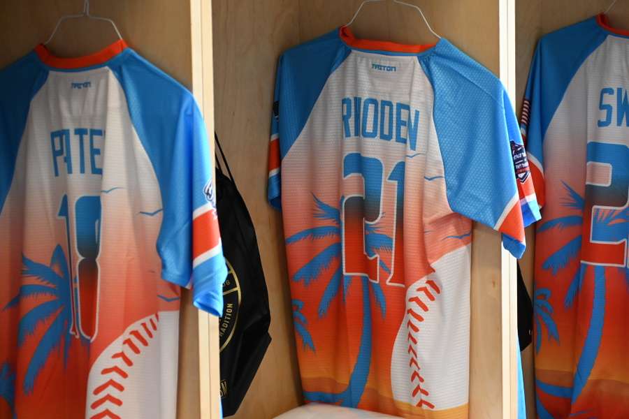 Three custom baseball jerseys hanging on hangers displaying player names and numbers in blue and orange.