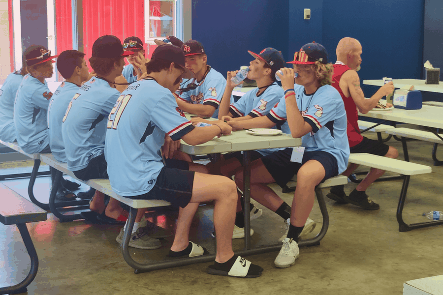 A youth baseball team in light blue uniforms sits together at a picnic table during a break.
