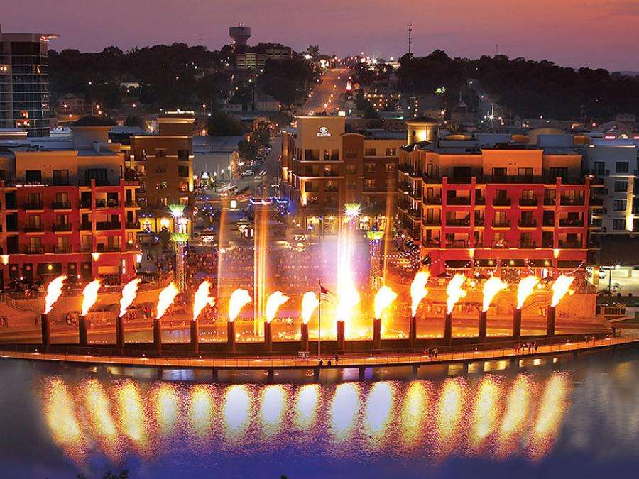 Waterfront plaza at dusk with illuminated flame torches reflecting in water and colorful buildings.