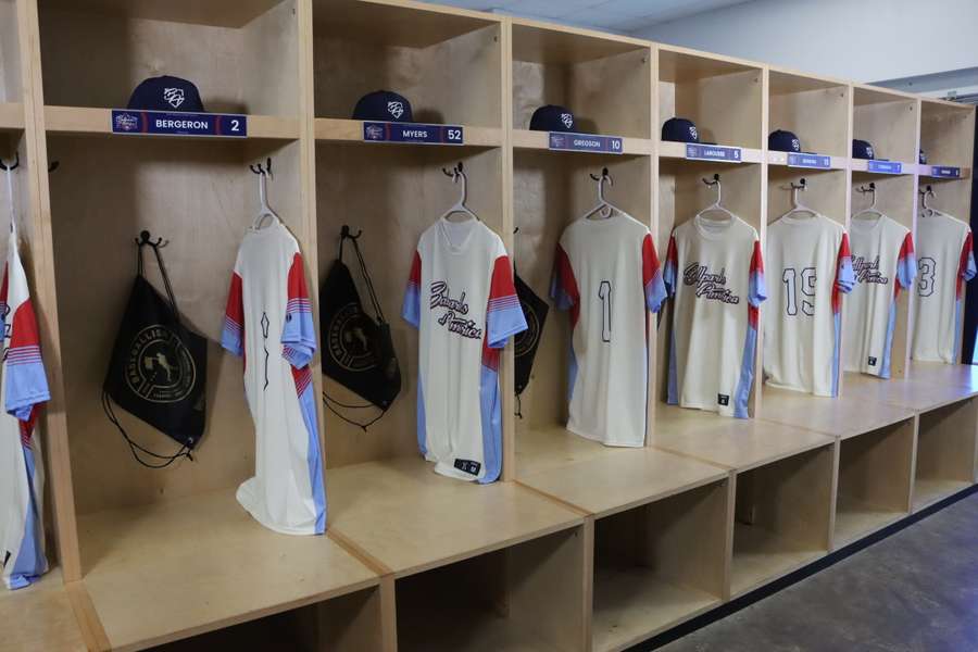 A sports locker room with wooden cubicles displaying numbered jerseys and team equipment on hangers.