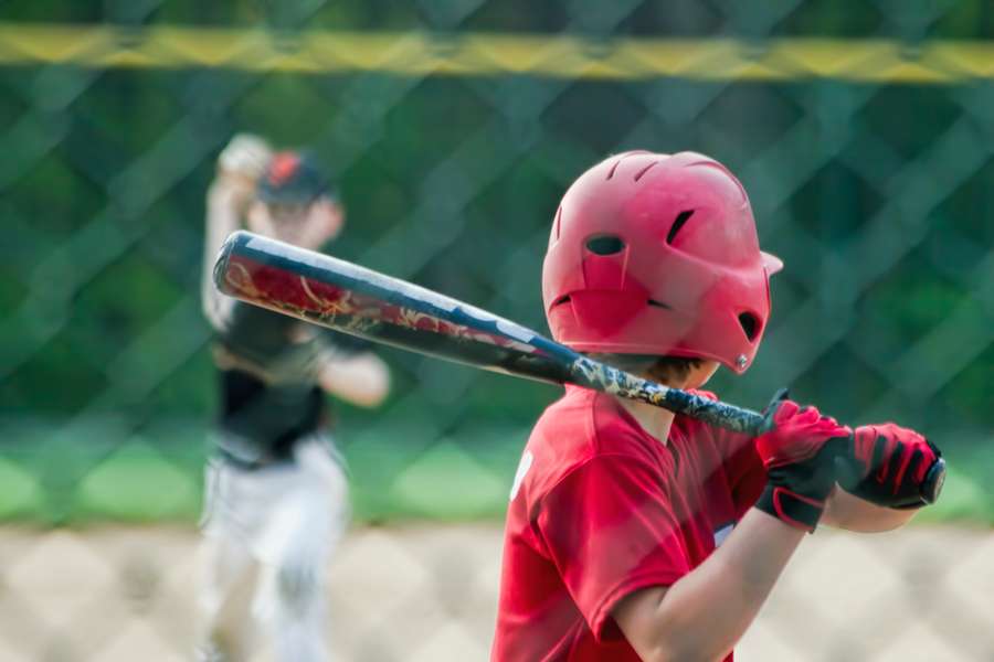 Young baseball player in red uniform holding bat with pitcher blurred in background.