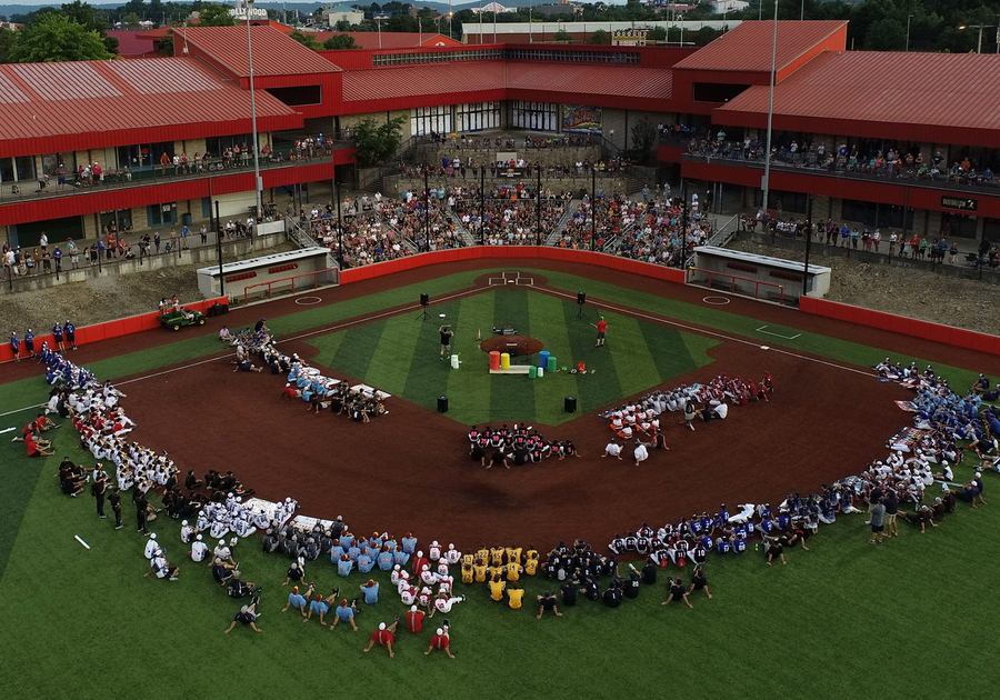 Multiple youth baseball teams lined up around a field during opening ceremony at a modern stadium.