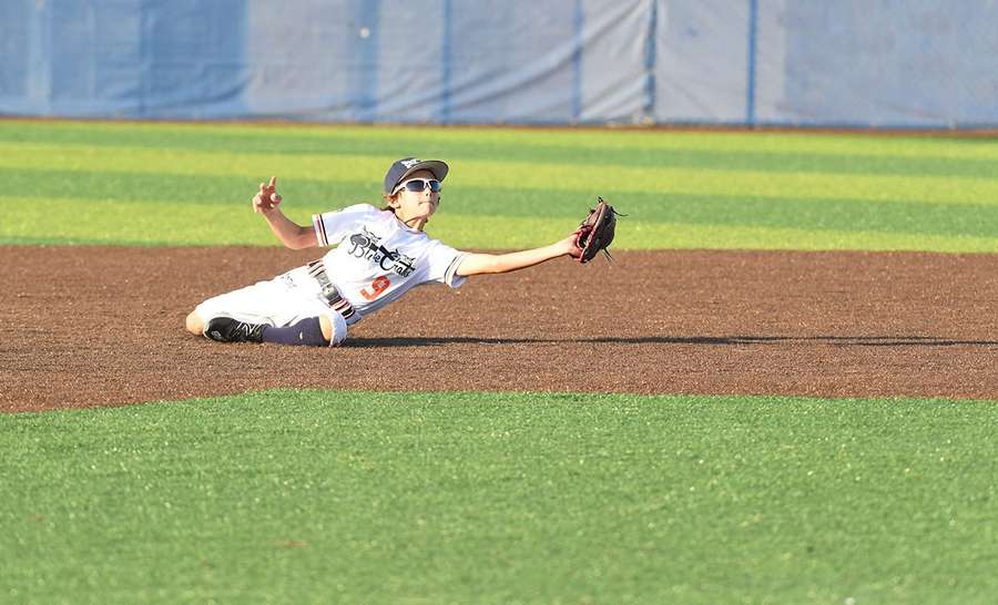 Young baseball player diving horizontally to catch a ball during outfield play at stadium.