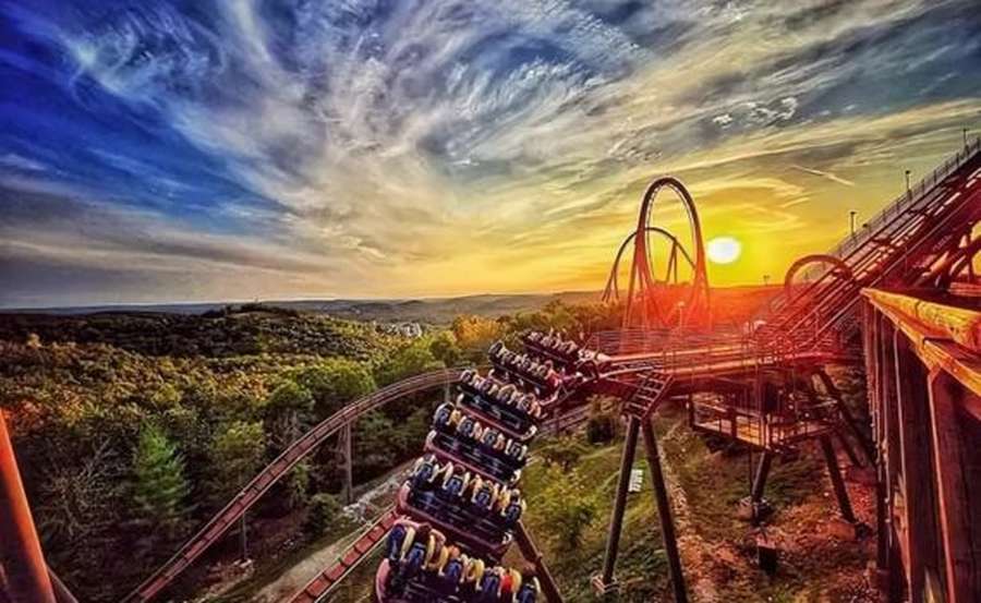 A roller coaster climbs through a forested landscape during a golden sunset with dramatic clouds.