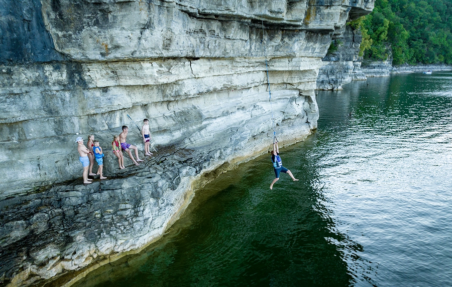 People cliff jumping and swimming in a clear green quarry lake surrounded by rocky limestone walls.