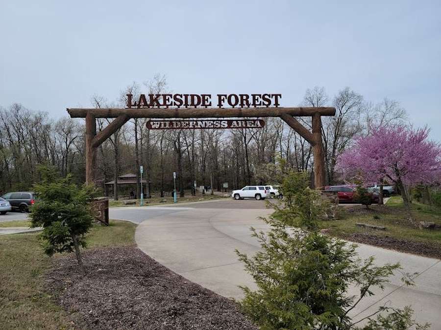 Large wooden entrance sign for Lakeside Forest Wilderness Area with parked vehicles and blooming trees.