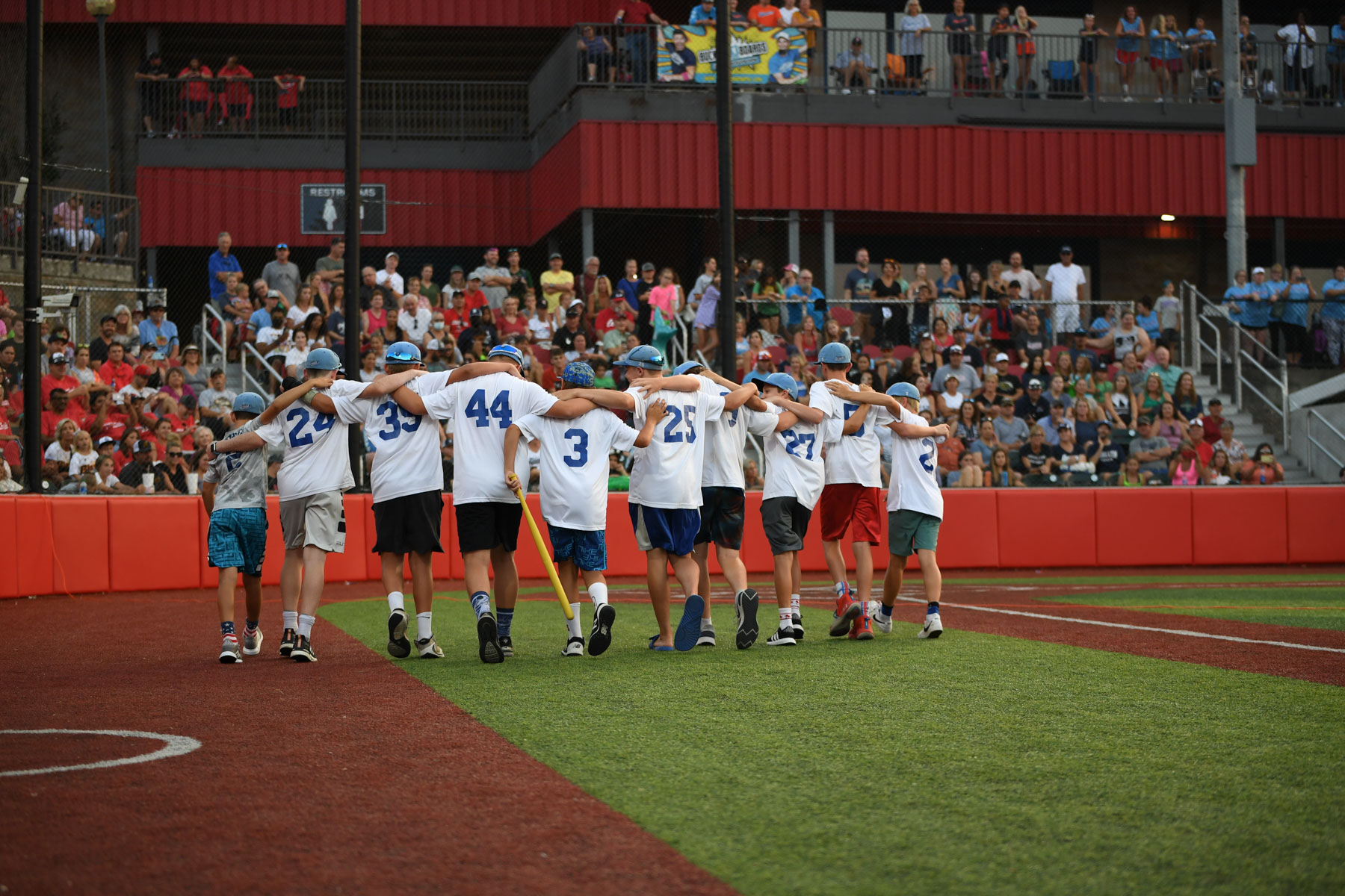 A team of youth travel baseball players have their arms around each other's shoulders as they walk off a baseball field. The players are wearing white jerseys with blue numbers.