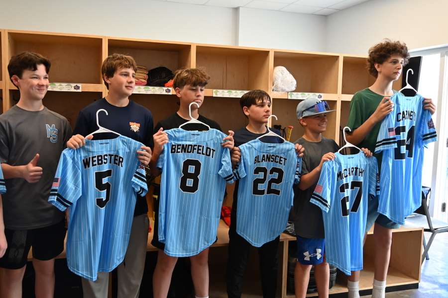 Six young baseball players displaying light blue pinstriped uniforms with their names and numbers on hangers.