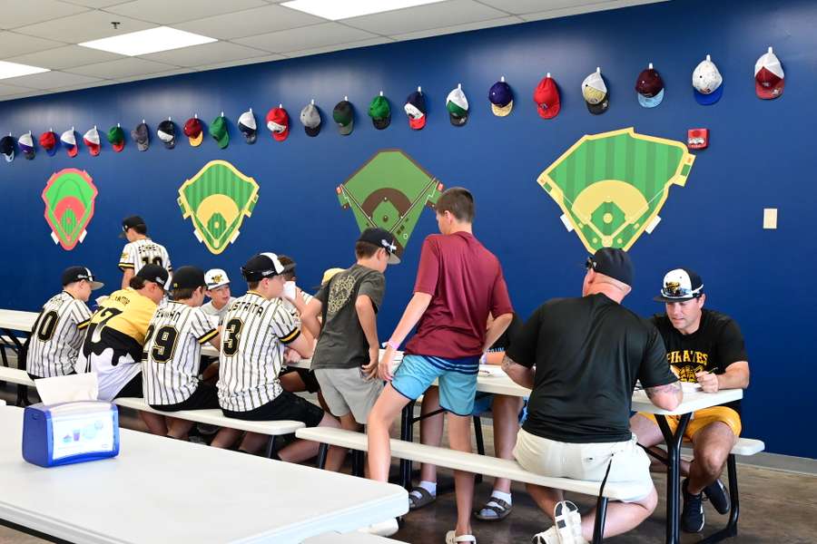 Young baseball players in Pirates uniforms sign autographs with coaches in dugout-style room.