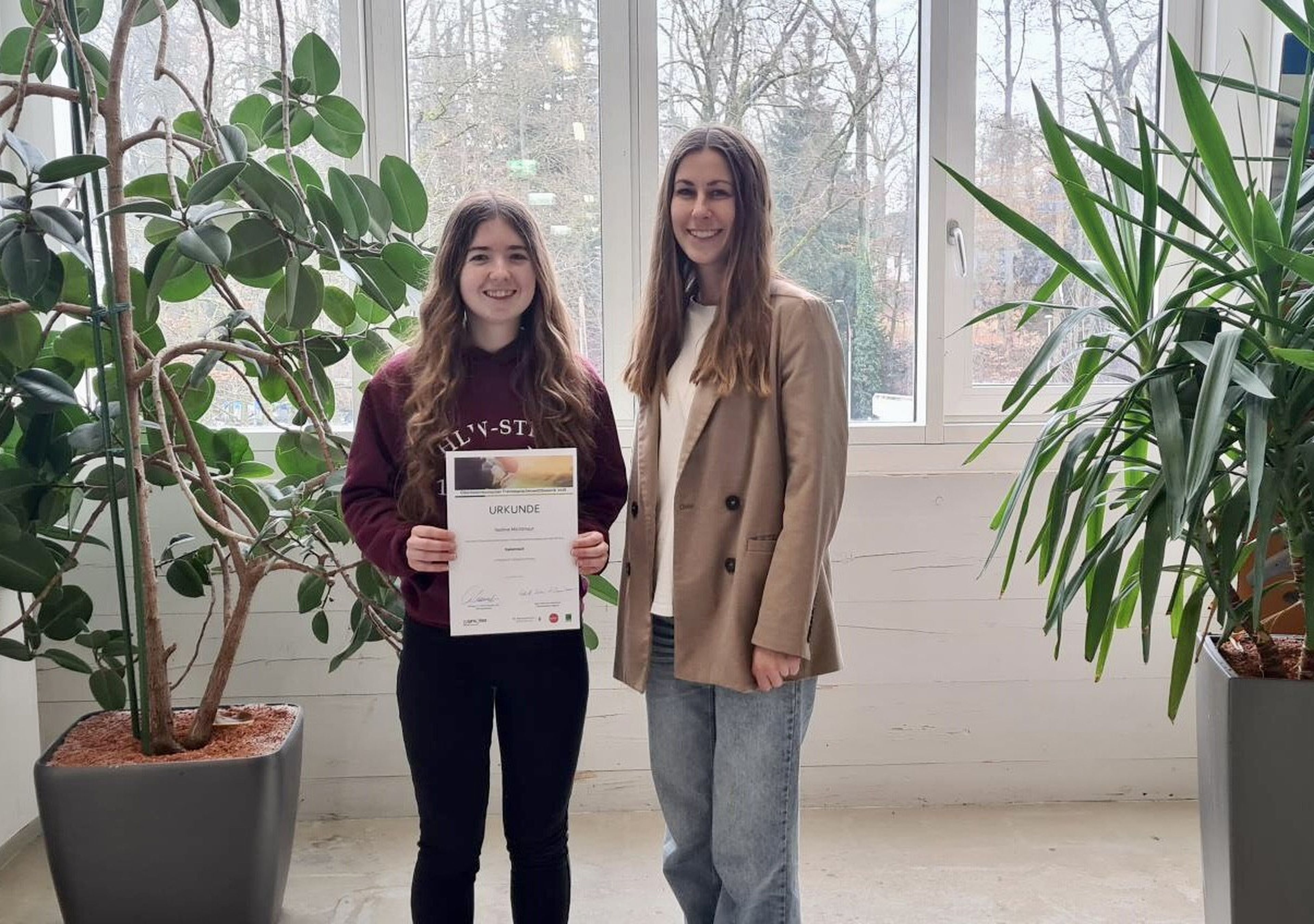 Two women smiling indoors; one holds a certificate. They're standing between two large potted plants near a window.