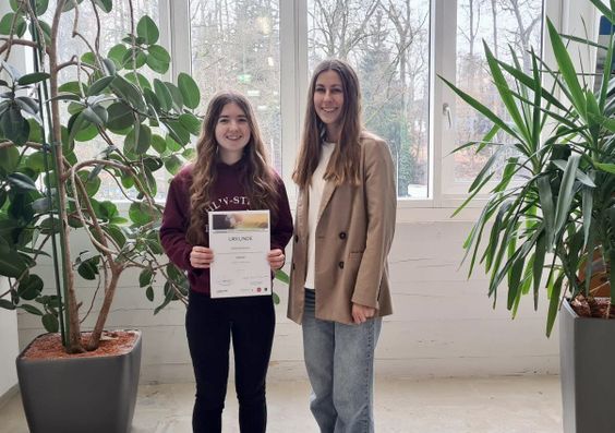 Two women smiling indoors; one holds a certificate. They're standing between two large potted plants near a window.