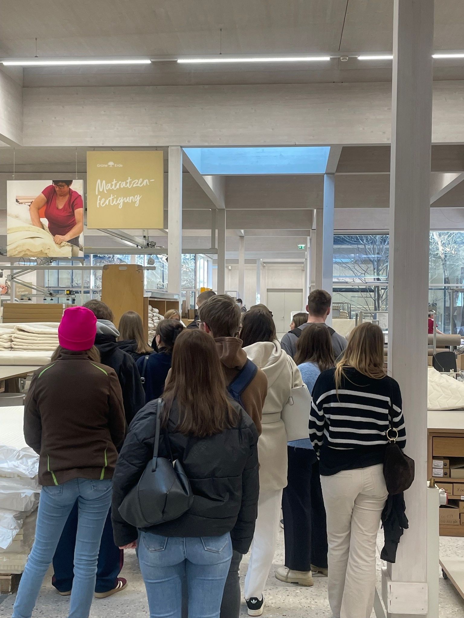 A group of people stand indoors, facing a display about mattress production. Bright lighting illuminates the space with large windows visible.