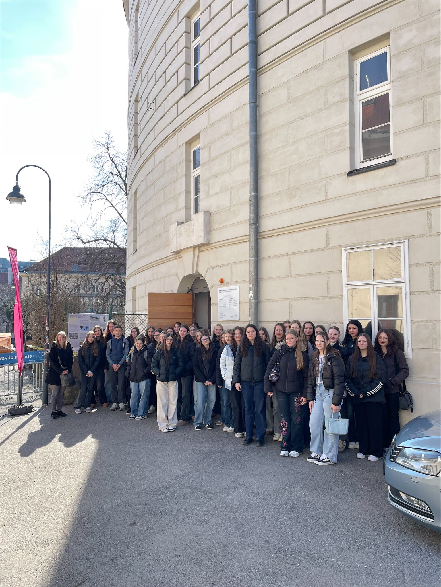A large group of people stand together outside a tall, round building on a sunny day.