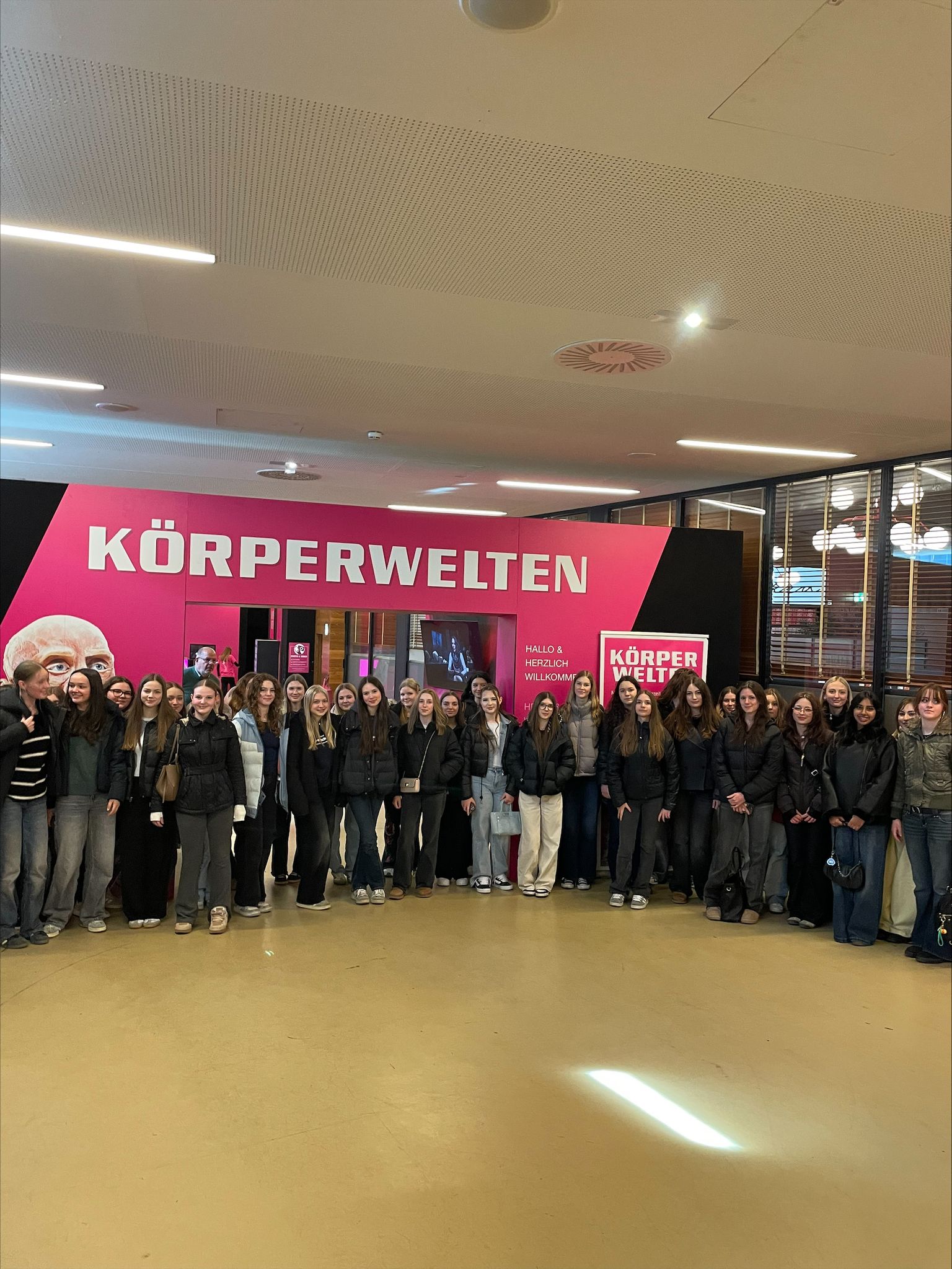 A group of people stands in front of a "Körperwelten" exhibition entrance, featuring a vibrant pink and black banner.